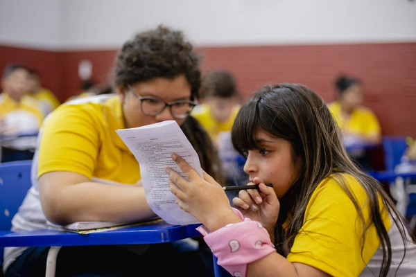 Dois alunos com camisas amarelas discutem um documento durante uma aula. Um aponta para o papel, enquanto o outro escuta atentamente, segurando uma caneta.