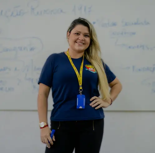 Uma mulher está em frente a um quadro branco, vestindo uma camisa azul e um cordão amarelo. Ela está segurando um marcador e sorrindo para a câmera, comemorando seus 30 anos. Texto e diagramas estão visíveis no quadro branco atrás dela.