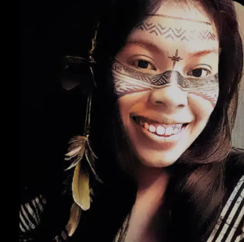 Uma mulher de cabelos longos sorri, rosto adornado com pintura cerimonial e acessório floral no cabelo, comemorando 30 anos.