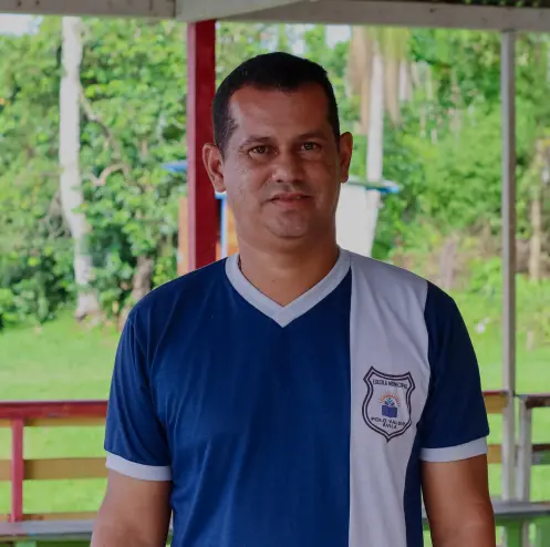 Um homem vestindo uma camiseta azul e branca com um emblema no peito, comemorando 30 anos, fica ao ar livre com vegetação e árvores ao fundo.