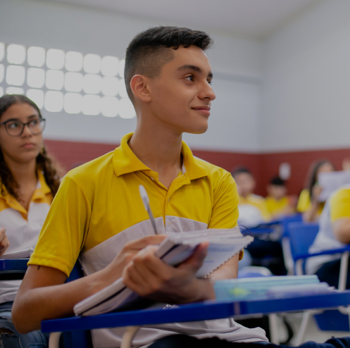 Um aluno vestindo uma camisa amarela e branca está sentado em uma mesa de sala de aula, segurando um caderno e uma caneta, olhando atentamente para a frente. Anos de aprendizagem são evidentes à medida que outros alunos são visíveis ao fundo.