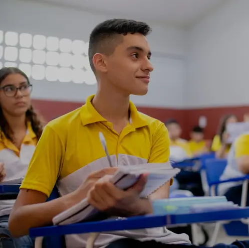 Um aluno vestindo uma camisa amarela e branca está sentado em uma mesa de sala de aula, segurando um caderno e uma caneta, olhando atentamente para a frente. Anos de aprendizagem são evidentes à medida que outros alunos são visíveis ao fundo.