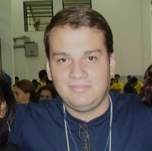 Uma pessoa de cabelos castanhos curtos, usando óculos e camisa azul, que aparenta ter cerca de 30 anos, está em uma sala com outras pessoas ao fundo.