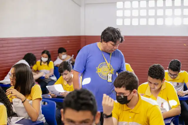 Um professor de camisa azul auxilia os alunos em uma sala de aula.