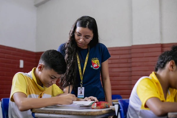 Uma professora de camisa azul auxilia um aluno escrevendo em uma carteira de sala de aula.