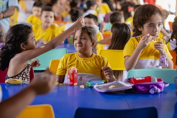 Na imagem, um grupo de crianças está sentado em mesas compridas em um refeitório escolar.