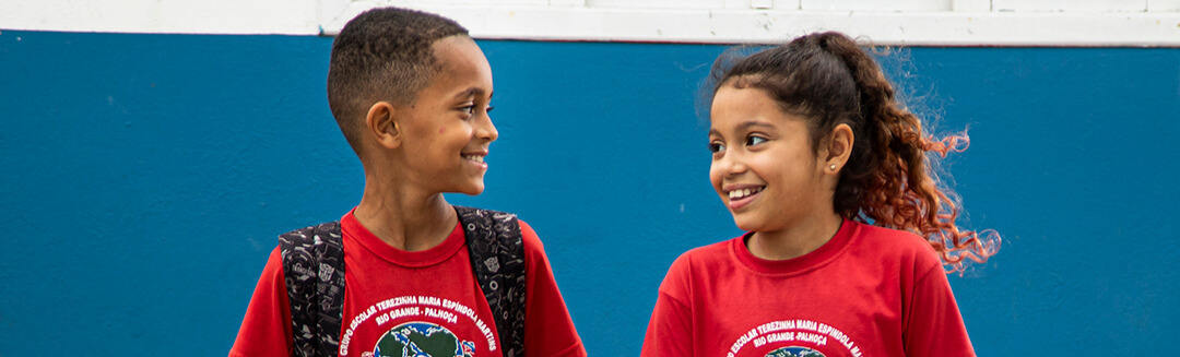 Duas crianças vestindo camisetas e mochilas vermelhas sorriem uma para a outra encostadas em uma parede azul e branca, refletindo o espírito do Marketing Relacionado à Causa Instituto Ayrton Senna.