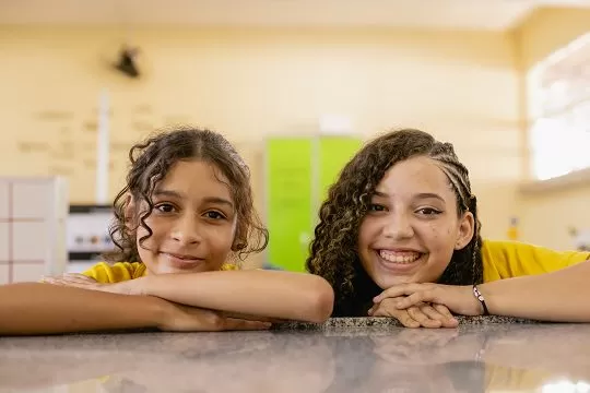Duas meninas apoiam os braços sobre uma bancada e sorriem para a câmera em uma sala iluminada e com paredes amarelas, celebrando o McDia Feliz para o Instituto Ayrton Senna.