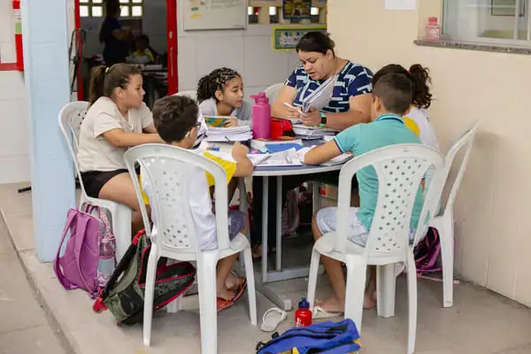 Um grupo de crianças está sentado ao redor de uma mesa com um professor, participando de uma sessão de estudo.