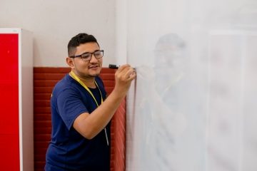 Pessoa de óculos e camisa azul, anotando um rascunho perspicaz com um marcador preto no quadro branco em uma sala de aula, em notícia sobre avaliação de carreira docente.