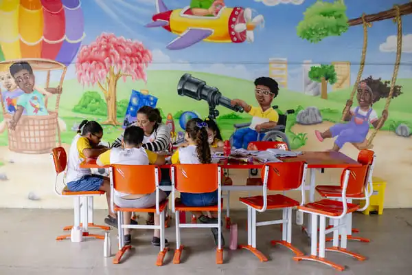 Crianças sentadas à mesa, envolvidas em atividades com professora.