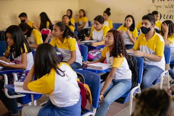 Alunos de uniforme amarelo e branco sentados em uma sala de aula, voltados atentamente para frente, alguns tomando notas enquanto outros observam.