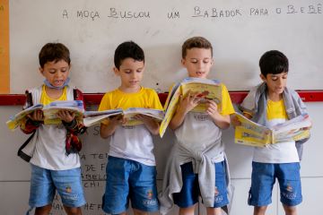 Quatro meninos com uniformes escolares iguais leem livros em voz alta em frente a um quadro branco com texto escrito à mão. Estudam em escola com boas notas no IDEB.