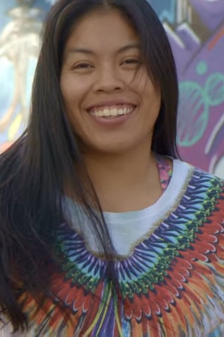 Uma pessoa com longos cabelos escuros está sorrindo e vestindo uma camisa colorida com estampas que lembram penas, comemorando 30 Anos de Legado em um cenário de grafite vibrante.