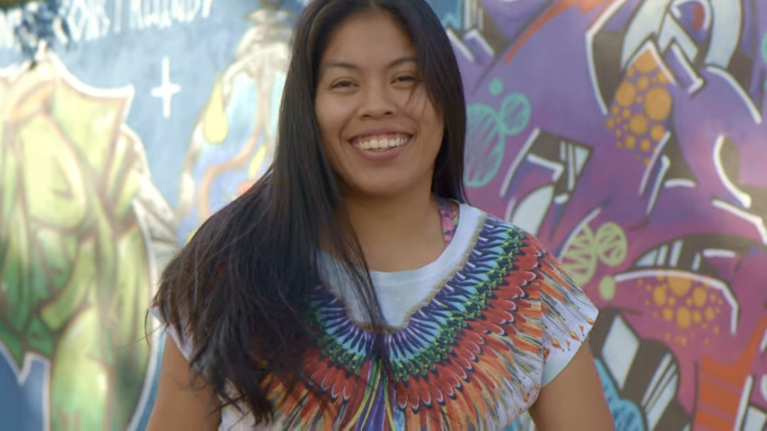 Uma pessoa com longos cabelos escuros sorri amplamente em frente a um muro de grafite colorido, vestindo uma camisa branca com um desenho de penas vibrantes em comemoração aos 30 Anos de Legado.