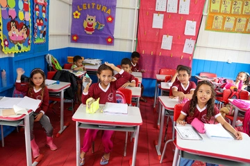 Uma sala de aula com crianças pequenas sentadas em carteiras, algumas levantando as mãos.