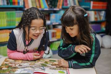 Duas meninas em idade de alfabetização leem um livro juntas em uma mesa em uma livraria ou biblioteca, com livros coloridos em prateleiras ao fundo.