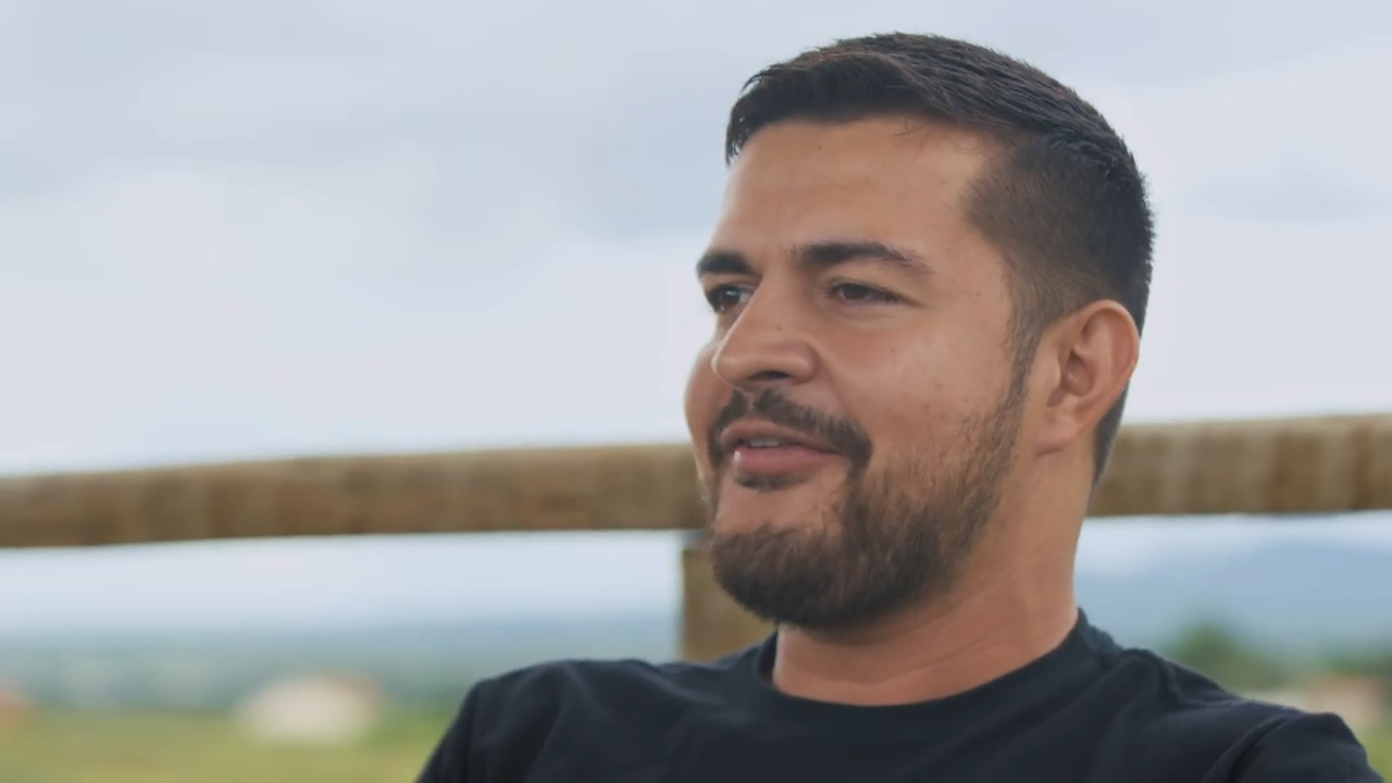 Um homem de cabelo curto e barba, vestindo uma camisa preta que celebra os "30 Anos de Legado", está sentado ao ar livre com um fundo desfocado.