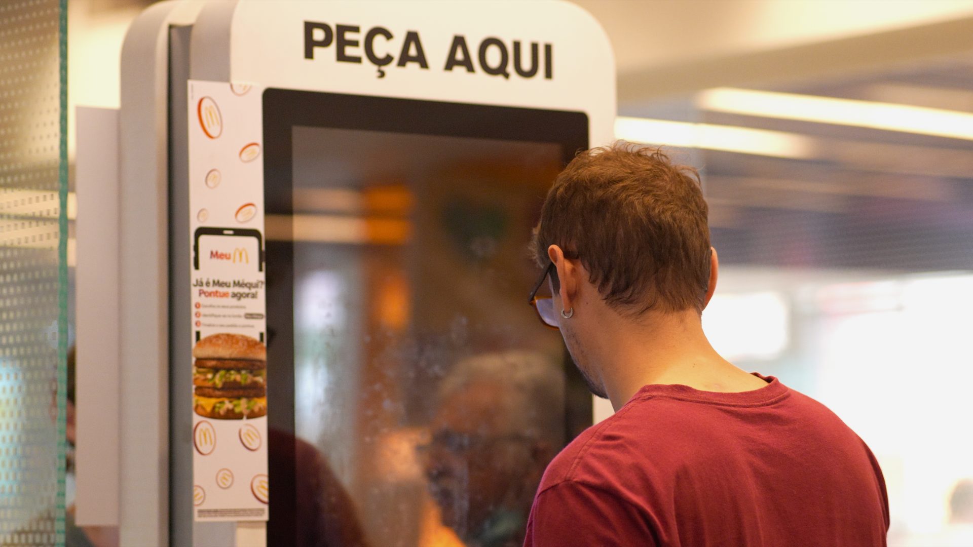 Um homem de óculos usa um quiosque de autoatendimento em um restaurante fast food. Acima da tela, o texto "PEÇA AQUI" o convida a fazer seu pedido, enquanto antecipa a emoção do McDia Feliz 2024.