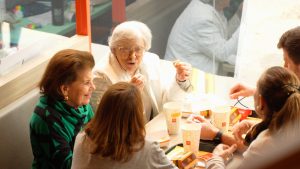 Um grupo de pessoas, incluindo duas mulheres idosas, sentam-se à mesa de um restaurante fast-food, compartilhando uma refeição e conversando durante o McDia Feliz 2024.