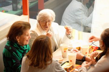 Um grupo de pessoas, incluindo duas mulheres idosas, sentam-se à mesa de um restaurante fast-food, compartilhando uma refeição e conversando durante o McDia Feliz 2024.