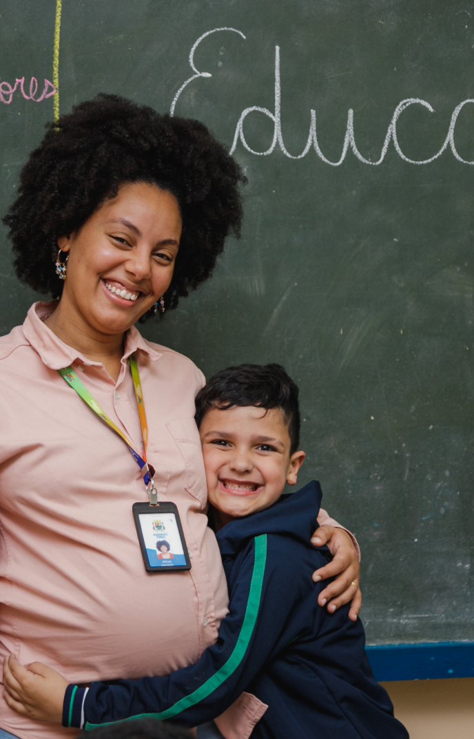 Um professor sorridente com um crachá posa com um jovem aluno em frente a um quadro negro com "Educ" escrito nele.