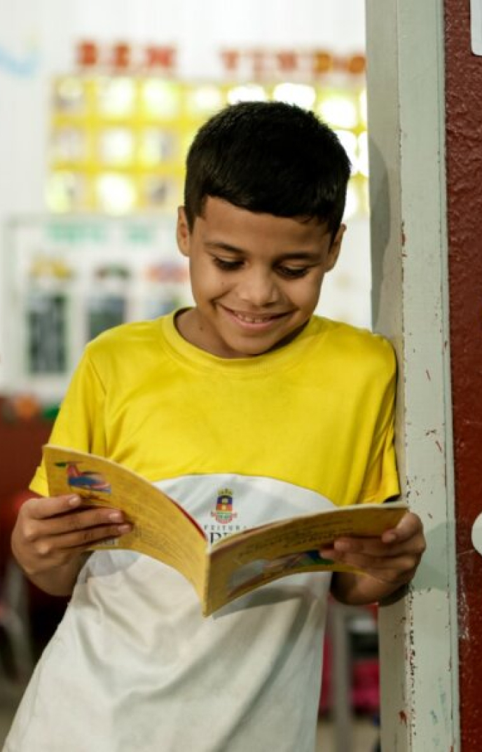 Um menino, comemorando "30 Anos de Legado" abraçando um legado de aprendizado, está encostado na parede, vestindo uma camisa amarela e branca, sorrindo enquanto mergulha em um livro em uma sala de aula.