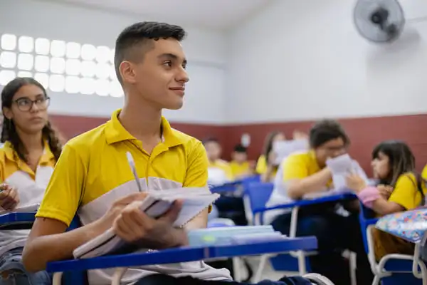 Um aluno em primeiro plano está segurando um caderno e sorrindo, enquanto outros no fundo estão envolvidos em atividades.