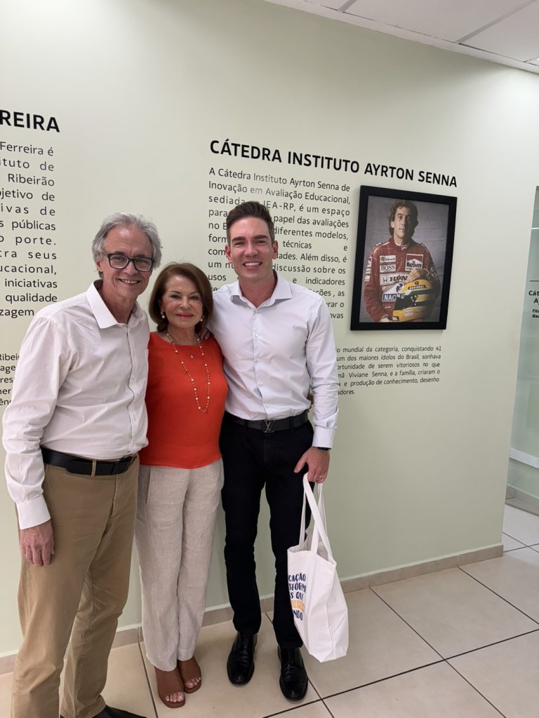 Três pessoas estão diante de uma parede adornada com texto e uma foto de alguém em traje de corrida, provavelmente simbolizando o Instituto Ayrton Senna. O homem à direita segura uma bolsa branca. Eles sorriem em seus trajes formais, incorporando o espírito dos estudios avançados da USP.