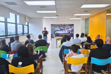 Uma mulher está na frente de uma sala, fazendo uma apresentação confiante para um público sentado, destacando sua parceria com o Instituto Ayrton Senna e o Itaú por meio do inovador programa Cartão.