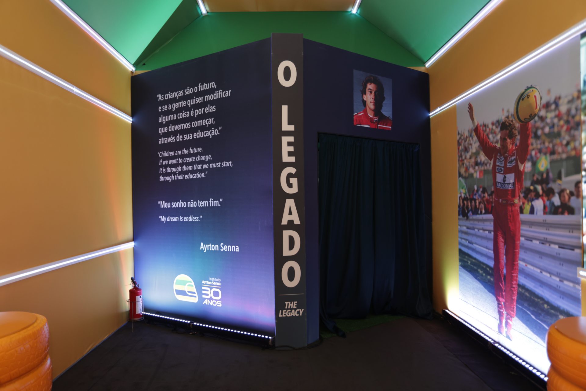 Uma exposição bem iluminada com uma homenagem a Ayrton Senna, apresentando sua imagem, citações e recordações, conecta-se ao espírito do McDia Feliz 2024 ao celebrar o legado de doação e excelência esportiva.