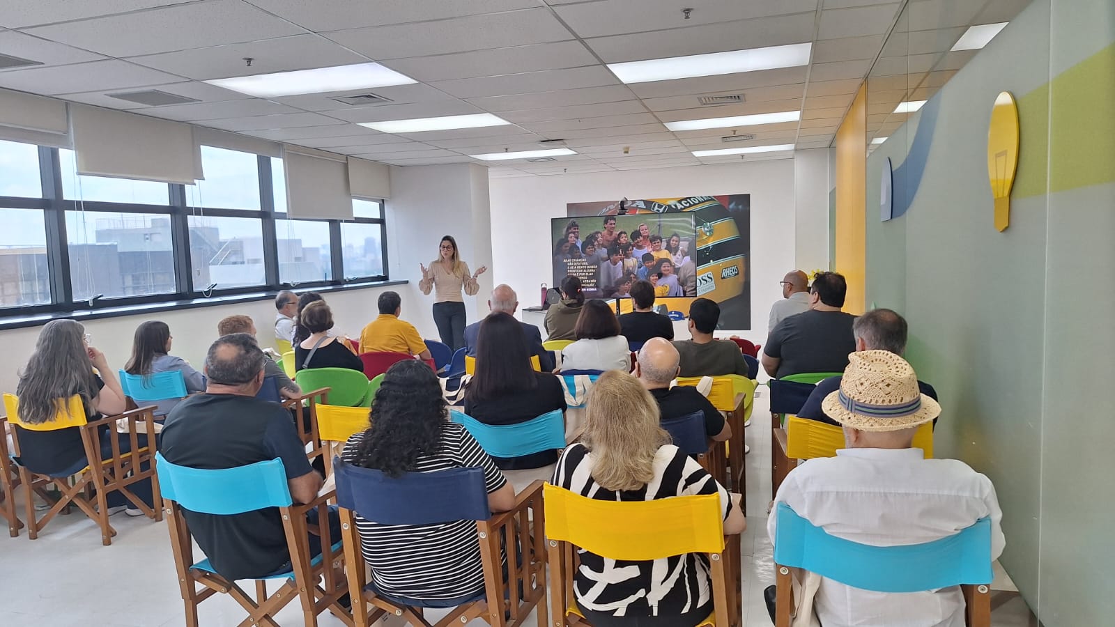 Um grupo de pessoas sentadas em cadeiras coloridas ouve atentamente um palestrante em uma sala iluminada, adornada com grandes janelas e decoração vibrante, talvez discutindo iniciativas apoiadas pelo Cartão Instituto Ayrton Senna Itaú.