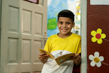 Uma criança sorridente está em pé na porta, segurando um livro aberto, cercada por recortes de flores coloridas. Graças ao PRONAC, essa cena alegre é enriquecida com a alegria de novas histórias e decorações.