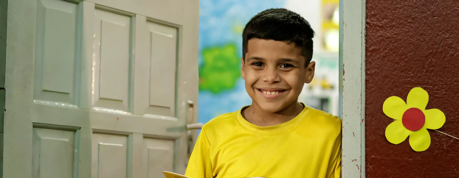 Um menino sorridente de camisa amarela está em frente a uma porta aberta decorada com uma flor amarela, simbolizando a generosidade da Doação iFood.