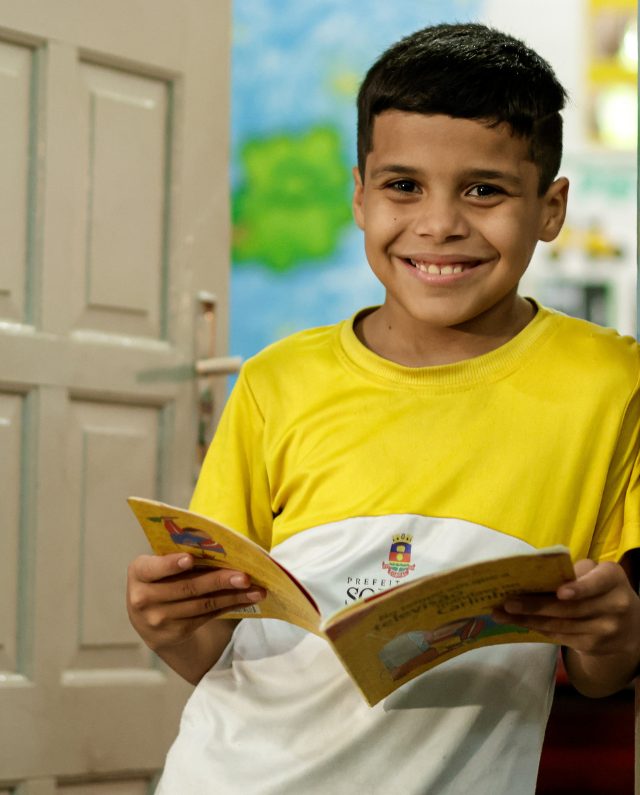 Um menino sorridente, de camisa amarela e branca, segura um livro aberto em uma sala bem iluminada, personificando a alegria daqueles que se beneficiam do Doação iFood, com uma porta parcialmente aberta ao fundo sugerindo infinitas possibilidades.