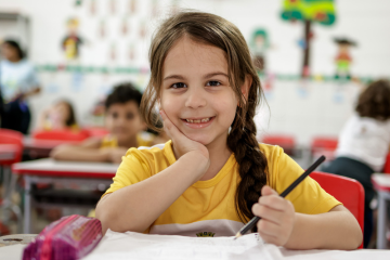 Uma jovem garota de camisa amarela está sentada em uma mesa de sala de aula, sorrindo e segurando um lápis com a mão esquerda na bochecha, personificando a alegria de aprender. Outras crianças e decorações de sala de aula são visíveis no vibrante ambiente educacional.