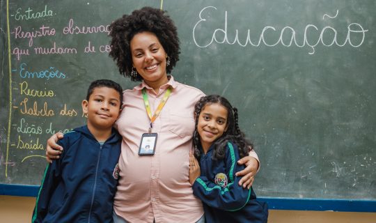 Uma professora está sorrindo entre duas crianças em frente a um quadro negro com "Educação" escrito nele. As crianças estão com os braços ao redor dela, personificando o espírito do compromisso do Instituto Ayrton Senna em promover a educação e o empoderamento.