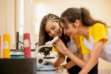 Em uma sala de aula do eduLab21, dois alunos vestidos com camisas amarelas e brancas se concentram intensamente enquanto olham através de um microscópio, explorando as maravilhas da ciência.