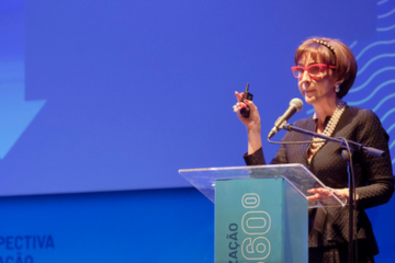 Uma pessoa com óculos vermelhos fala apaixonadamente em um pódio em um palco azul, segurando um dispositivo em uma mão, incorporando o espírito de defesa e mobilização.