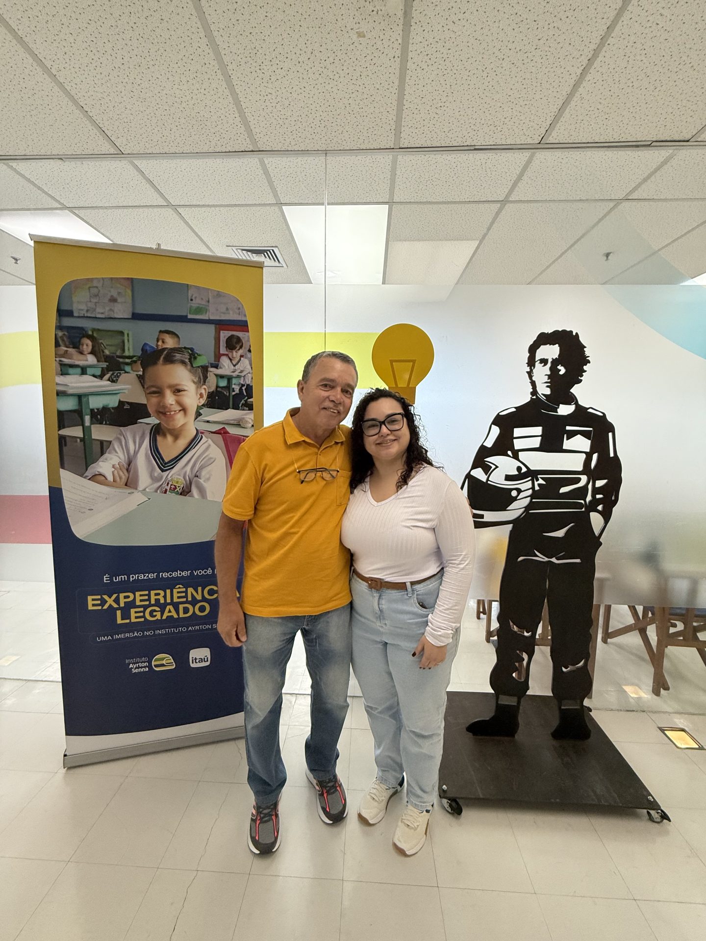 Duas pessoas estão juntas em frente a um display com um banner promovendo o Cartão Instituto Ayrton Senna Itaú, ao lado de um jogador de futebol.