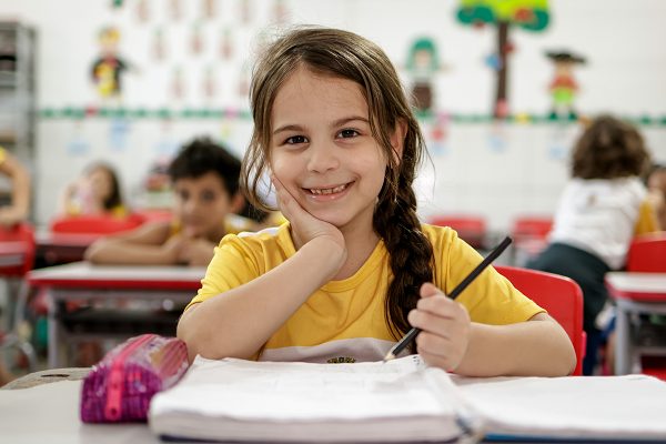 Uma jovem com cabelos trançados sorri para sua mesa em uma sala de aula, segurando um lápis e sentada em frente a um caderno, cercada por alunos. Decorações de sala de aula e placas destacando doações trazem um calor extra ao fundo.