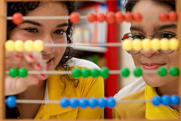 Dois estudantes de camisa amarela usam um ábaco com contas vibrantes vermelhas, amarelas, verdes e azuis. Um ajusta uma conta enquanto o outro observa atentamente, aprendendo o valor da contagem além dos números — como o impacto das doações via lei rouanet