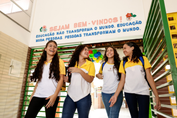 Quatro estudantes em uniformes amarelos e brancos caminham juntos sob uma placa de boas-vindas em português na entrada de uma escola, criando um cenário que lembra a capa de uma revista educacional.