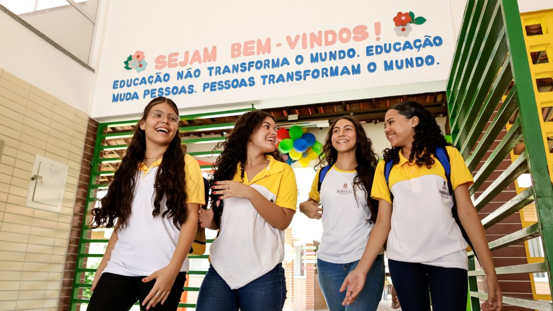 Quatro estudantes em uniformes escolares passeiam alegremente sob uma placa com texto motivacional sobre educação em português, personificando o espírito de aprendizado encontrado na Revista Educação.