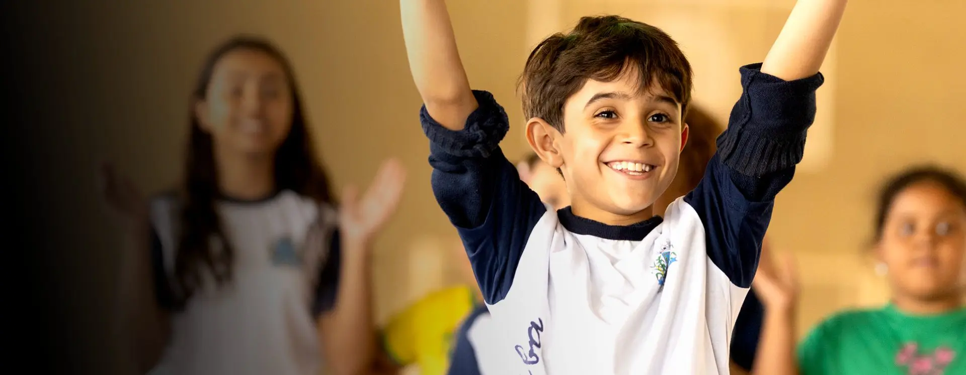 Um menino sorridente levanta orgulhosamente os braços em frente a um fundo desfocado de outras crianças, todas com uniformes escolares semelhantes, personificando o espírito vibrante promovido pelo Instituto Ayrton Senna.
