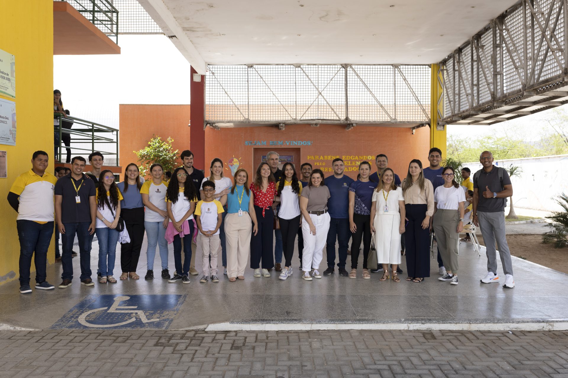 Um grupo de pessoas posa para uma foto do lado de fora da entrada de um prédio, perto de um símbolo azul de acessibilidade para cadeira de rodas pintado no chão, representando a coalizão Educação Já.