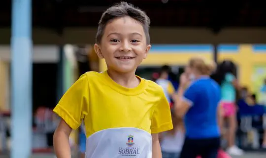 Um menino sorridente vestindo uma camisa amarela e branca do uniforme do Instituto Ayrton Senna com a inscrição "Prefeitura de Sobral" está ao ar livre; outras pessoas aparecem desfocadas no fundo.