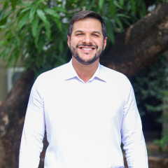 Um homem com cabelo escuro curto e barba, vestindo uma camisa branca, está ao ar livre em frente a uma árvore, sorrindo para a câmera como se estivesse posando para um rascunho automático.
