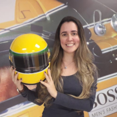 Mulher de cabelos longos sorri enquanto segura um capacete de corrida amarelo, em frente a anúncios de carros e moda, capturando um momento que parece um vibrante Rascunho automático.