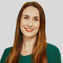 Cíntia Azone, uma mulher com longos cabelos castanhos, vestindo um top verde, sorri para a câmera contra um fundo cinza claro.